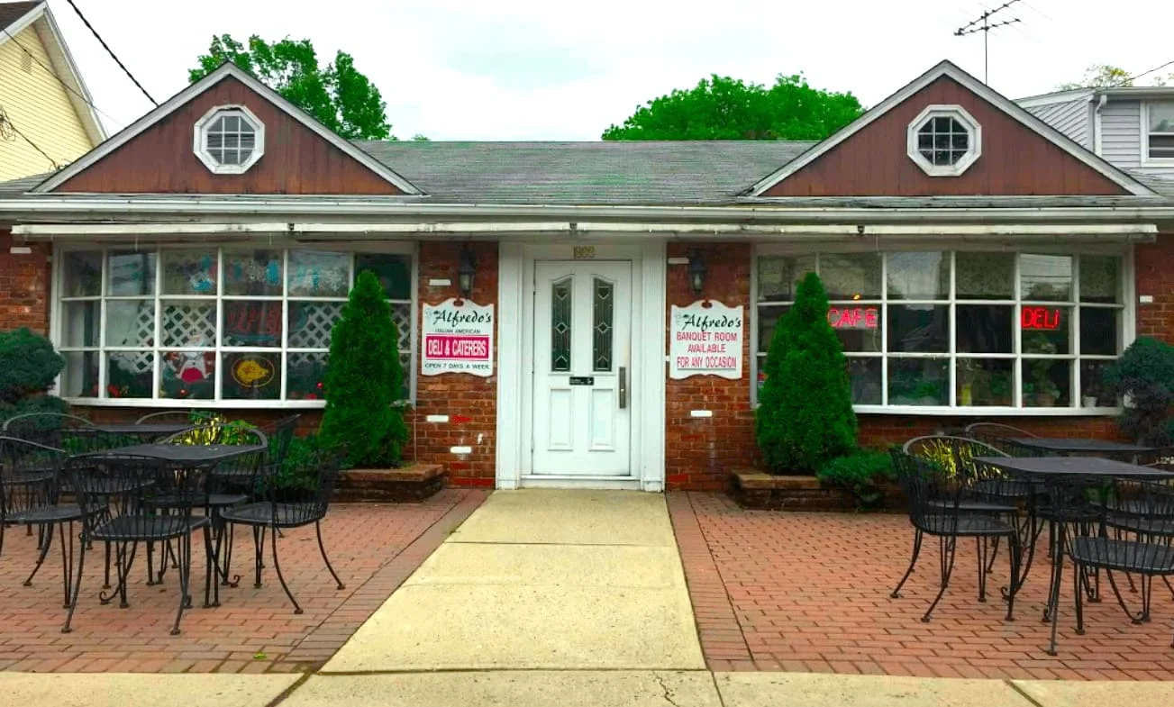 The deli storefront: a brick front with a white door, window signs for the deli and the banquet room, and patio tables on both sides