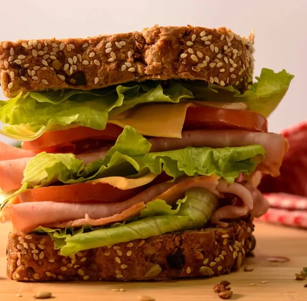 A tall sandwich of sliced ham, cheese, tomato and lettuce on seeded bread, with a red gingham towel behind it
